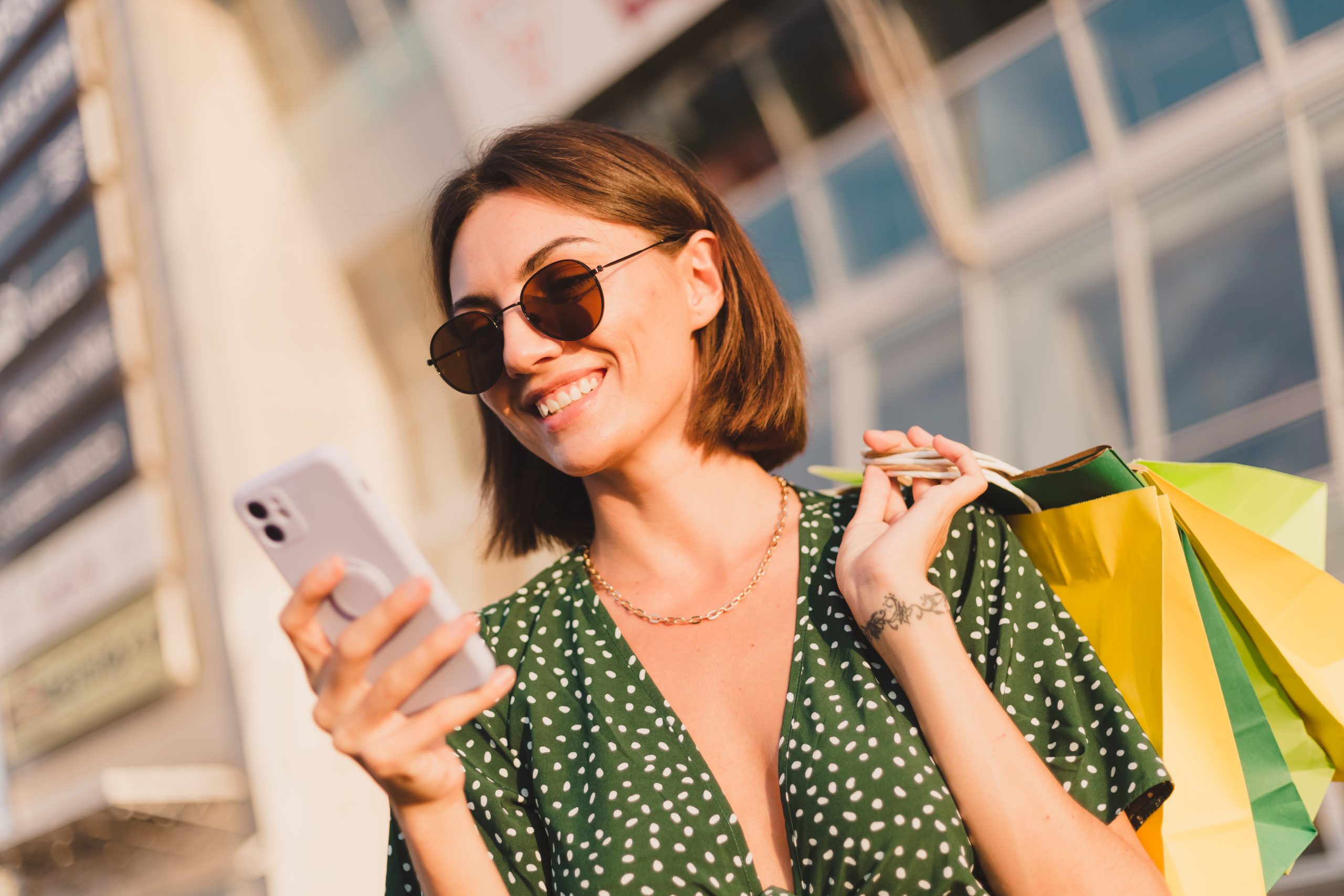 Woman at sunset with colorful shopping bags and parking lot by shopping mall happy with mobile phone